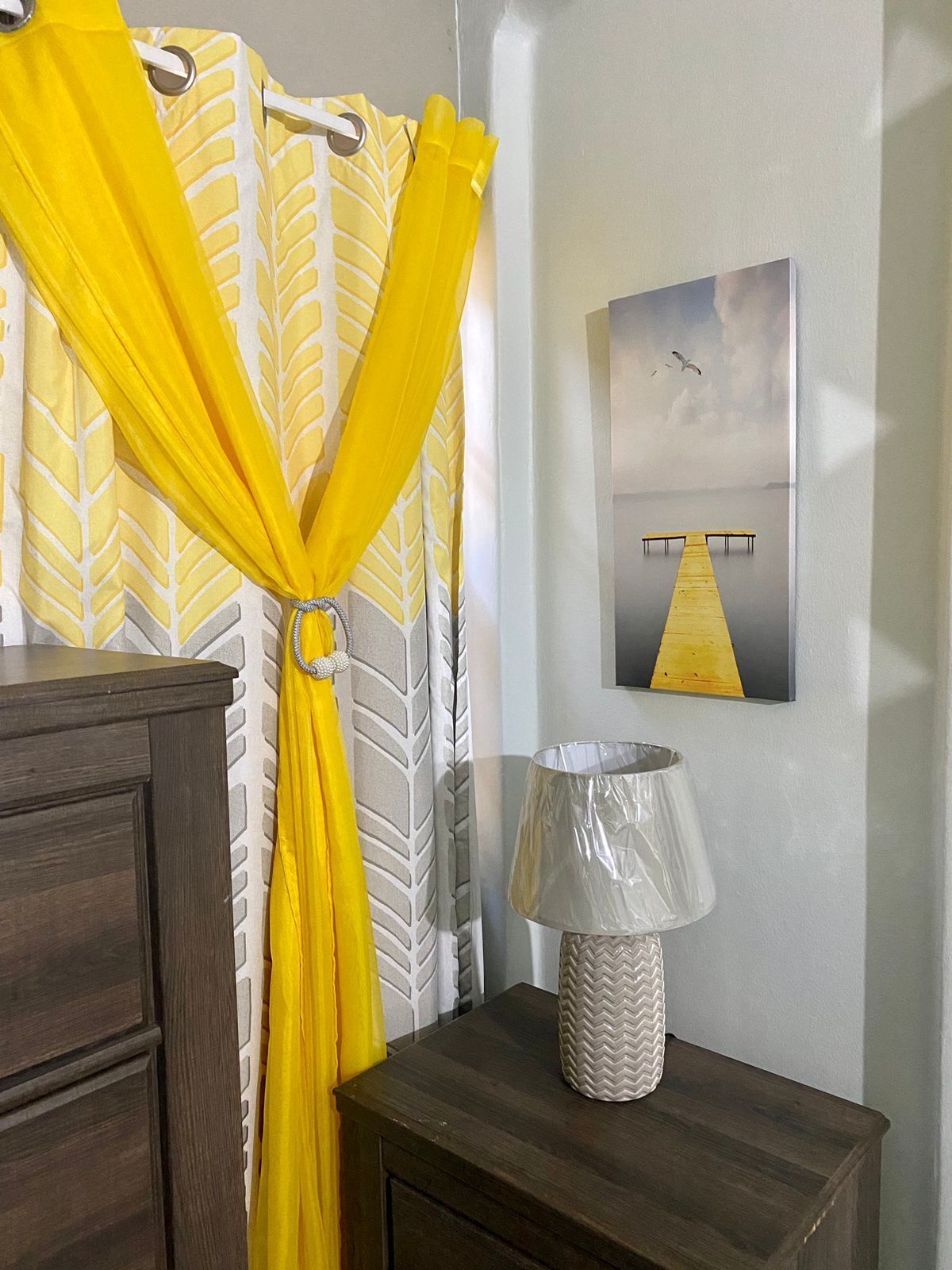 A room with a wooden dresser, a lamp with a chevron-patterned lampshade, a yellow curtain with a leaf pattern, and a painting of a dock with a seagull on the wall.