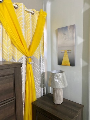 A room with a wooden dresser, a lamp with a chevron-patterned lampshade, a yellow curtain with a leaf pattern, and a painting of a dock with a seagull on the wall. - Luxury Home Hughenden Kingston (Kingston)