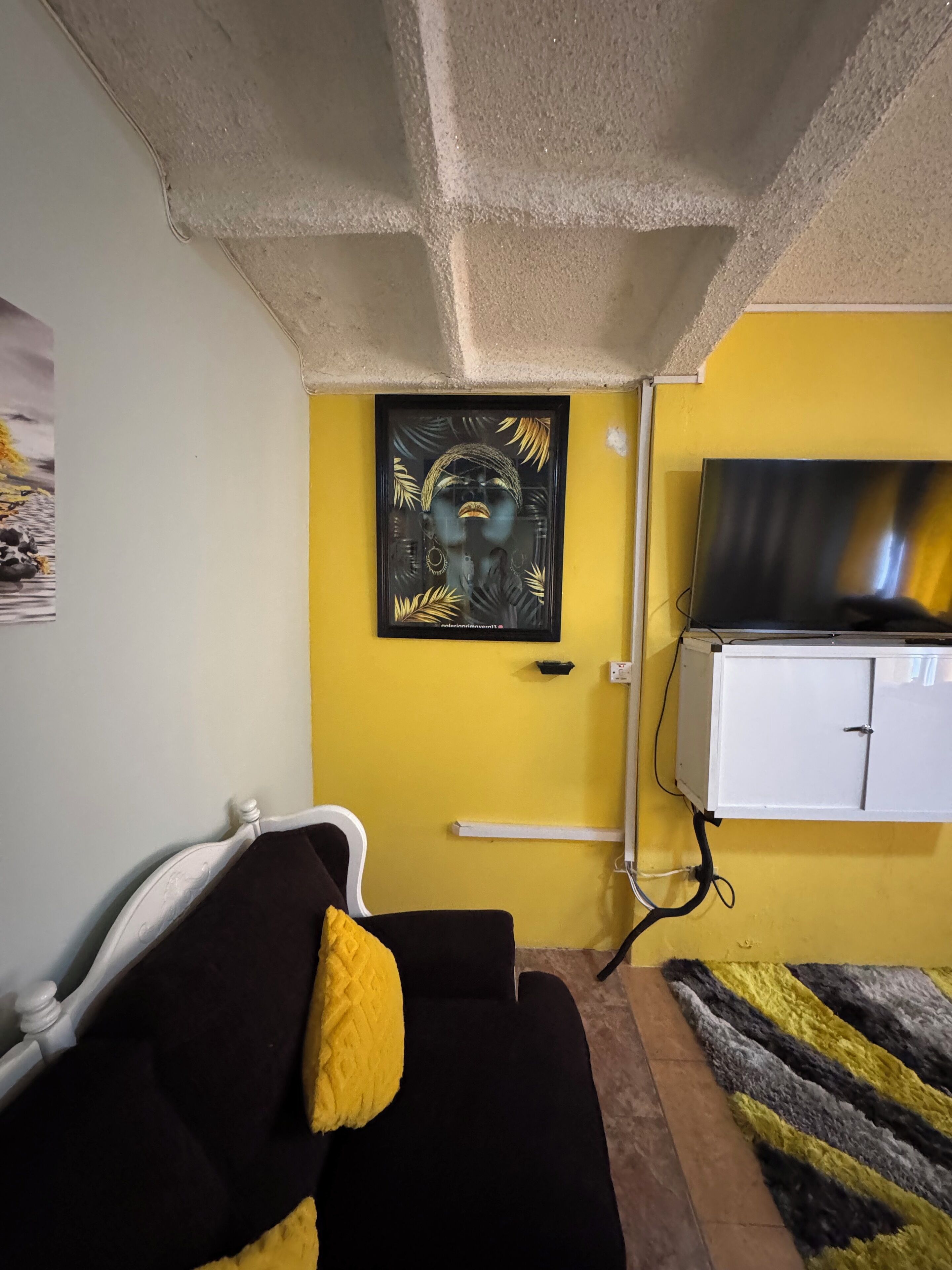 A room with a yellow wall, a black and white sofa with a yellow pillow, a TV on a white cabinet, and a framed artwork above the sofa.