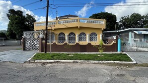 Un edificio de dos pisos con fachada amarilla, entrada protegida por una reja y un pequeño jardín delantero.
