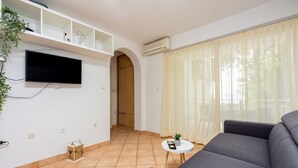Een moderne woonkamer met een grijze bank, een flatscreen televisie aan de muur, een klein rond tafeltje met een plant en een groot raam met lichte gordijnen.