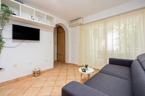 Ein modernes Wohnzimmer mit grauem Sofa, einem flachbildschirmfernseher an der Wand, einem kleinen runden Tisch mit Pflanze und einem großen Fenster mit leichtem Vorhang.