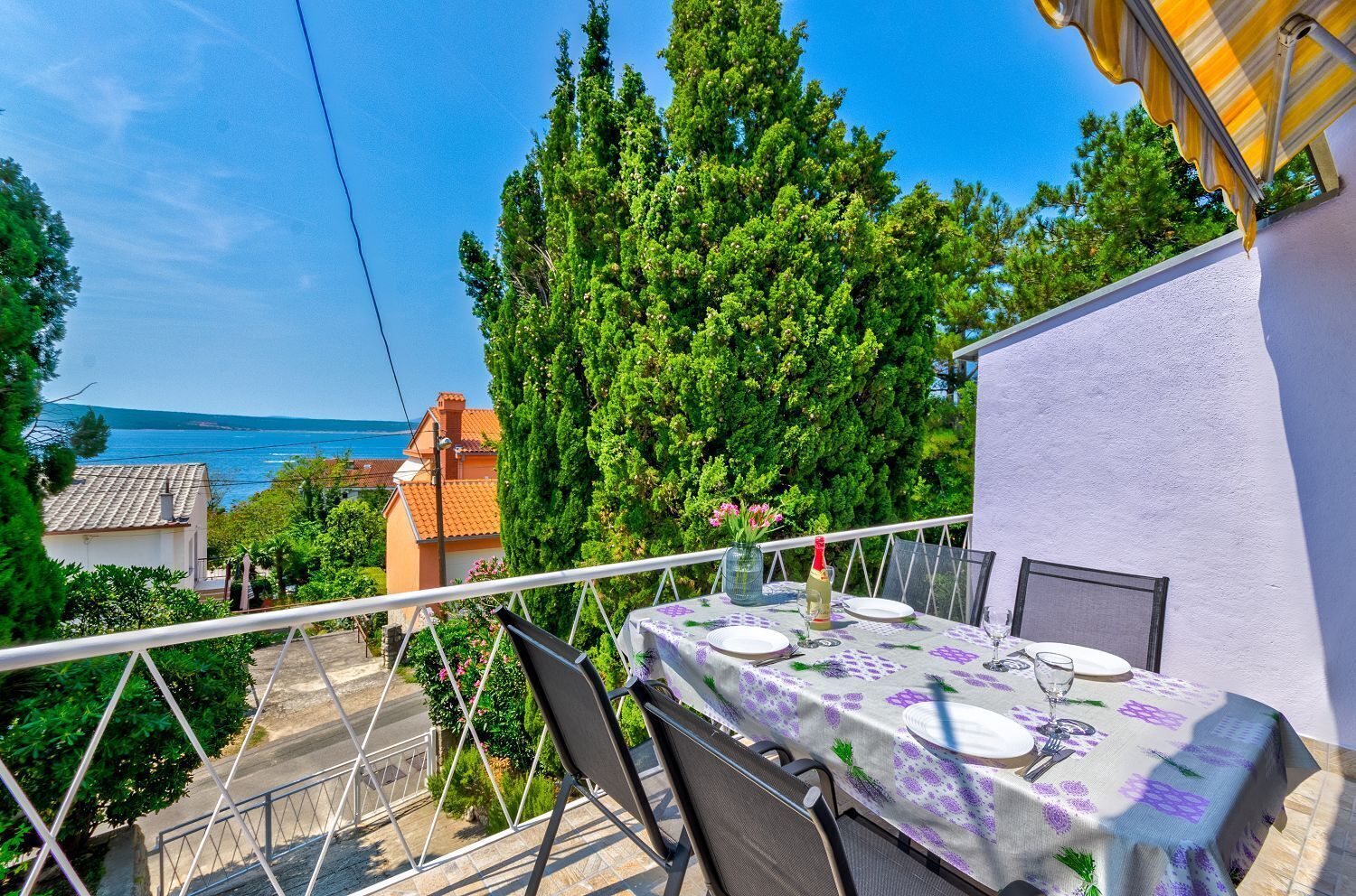An outdoor dining area with a table set for two, overlooking a scenic view of the sea and lush greenery.