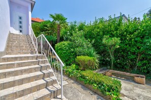 Eine Treppe aus Stein führt zu einer weißen Tür, umgeben von üppigem Grün und einem klaren Himmel.