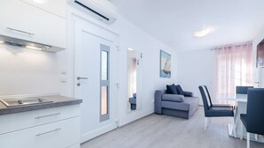 Ett modernt, väl upplyst rum med ett litet kök, en grå soffa, ett matbord med stolar och ett stort fönster med gardiner.