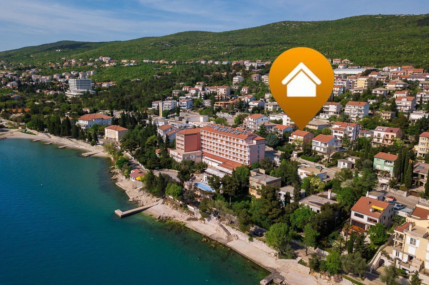 An aerial view of a coastal town with a prominent hotel featuring multiple floors and a red roof, surrounded by residential buildings and a coastline.