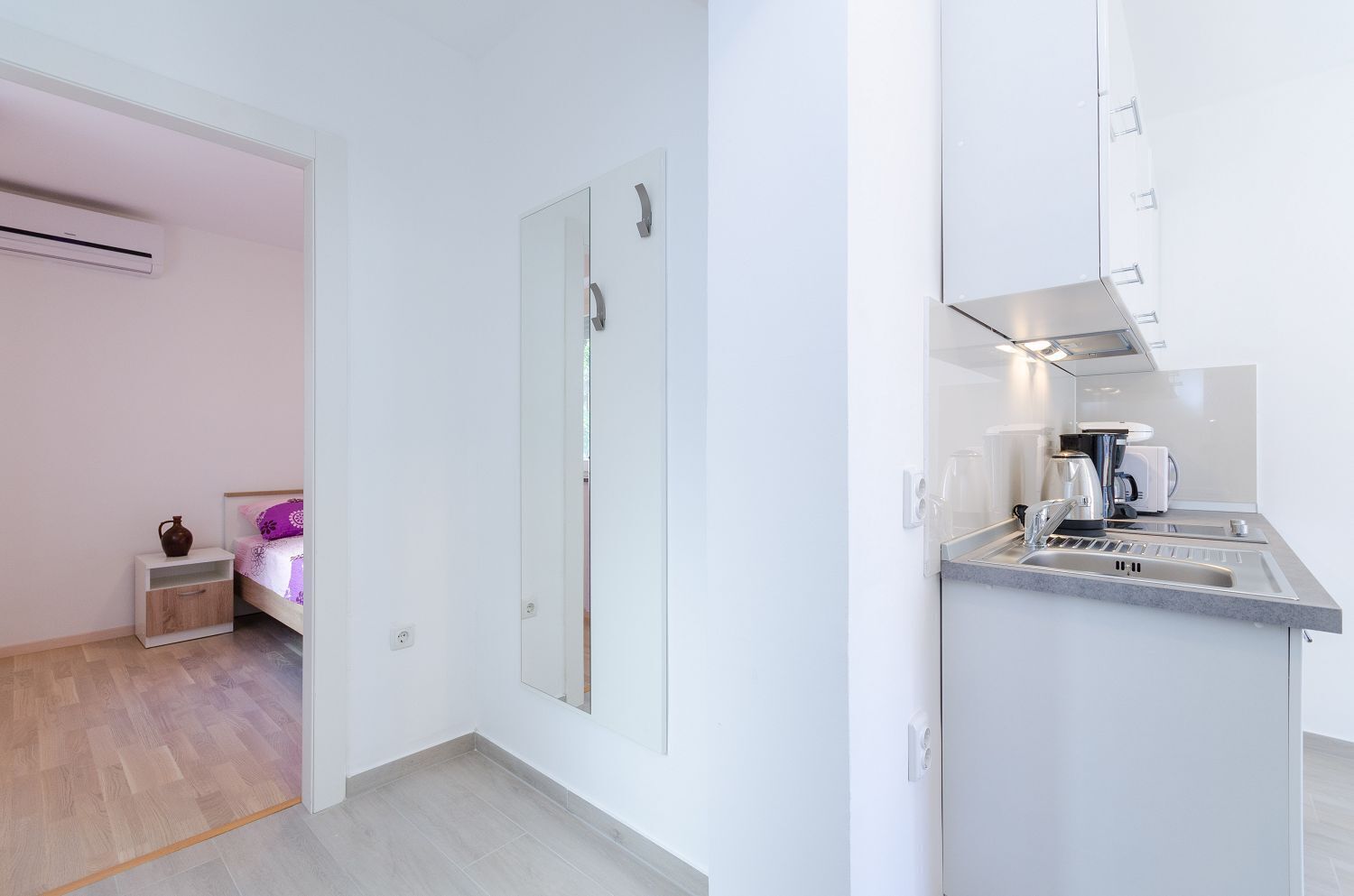 A modern, minimalist room with a bed, bedside table, and a small nightstand. A large mirror is mounted on the wall, and a kitchen area with a sink and countertop is visible.