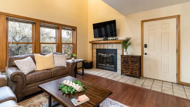 A living room with a fireplace, a television mounted above it, and a door to the right. - Rustic In-Town Retreat | Pool | Hot Tub (Durango)
