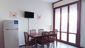 A compact living space with a dining table, chairs, a refrigerator, a television, and a sliding glass door to a balcony. - Cozy Flat With Pool Near the Beach (Jesolo)