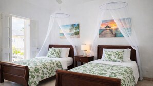 Une chambre avec deux lits en bois, chacun doté d’un baldaquin et de draps à motifs floraux. Une table de chevet avec une lampe se trouve entre les lits, et un tableau représentant un coucher de soleil est accroché au mur.