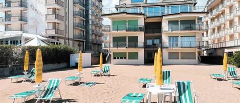 A hotel with multiple balconies, a sandy beach area with sun loungers and umbrellas, and a clear blue sky.