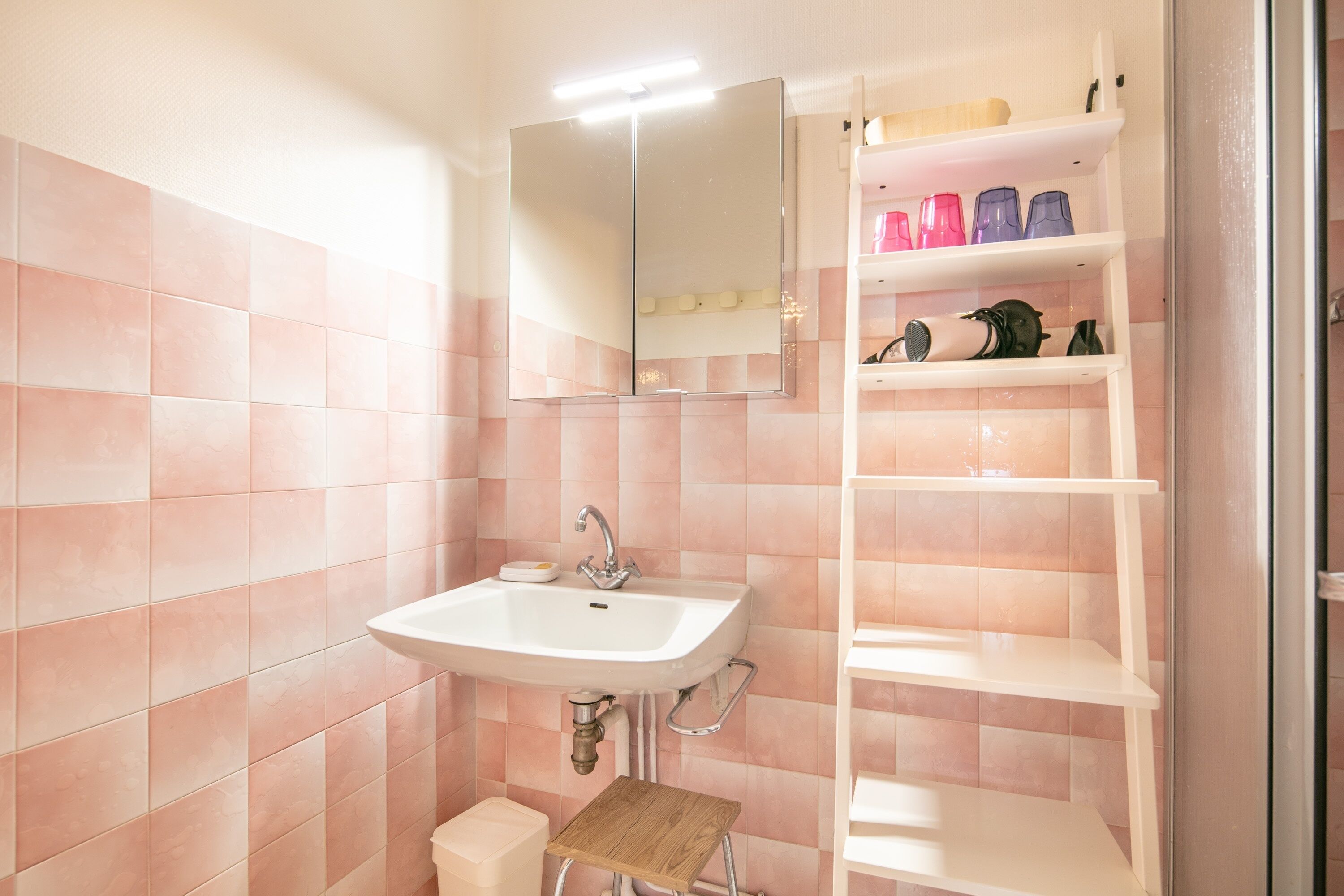 Um banheiro com azulejos rosa, pia branca, espelho e prateleira com artigos de higiene pessoal.