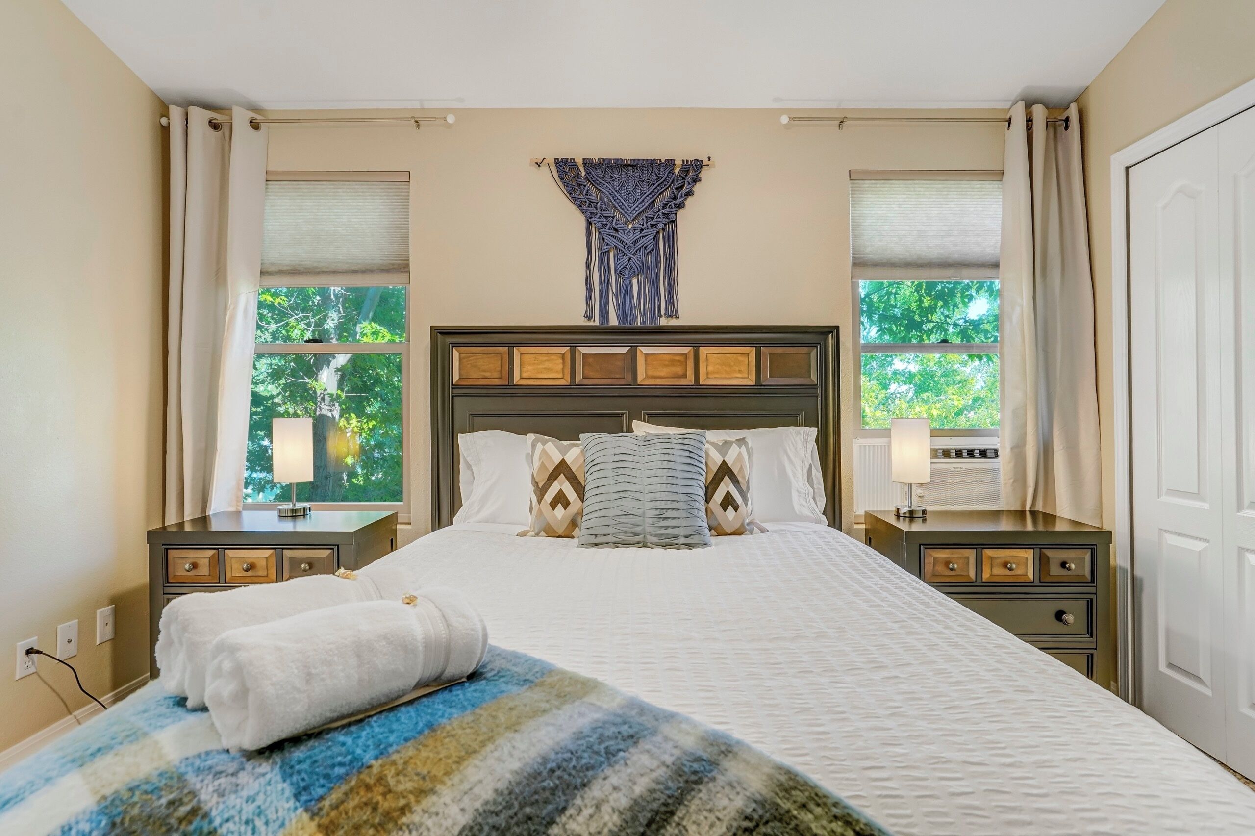A hotel room with a bed, two bedside tables, a lamp, a window with a view of trees, and a wall-mounted artwork.