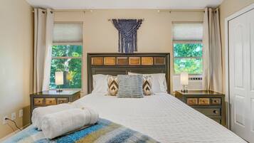 A hotel room with a bed, two bedside tables, a lamp, a window with a view of trees, and a wall-mounted artwork.