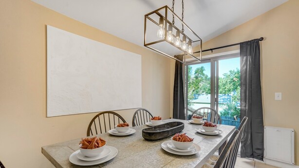 A dining area with a large table, chairs, a modern chandelier, and a view through the window.