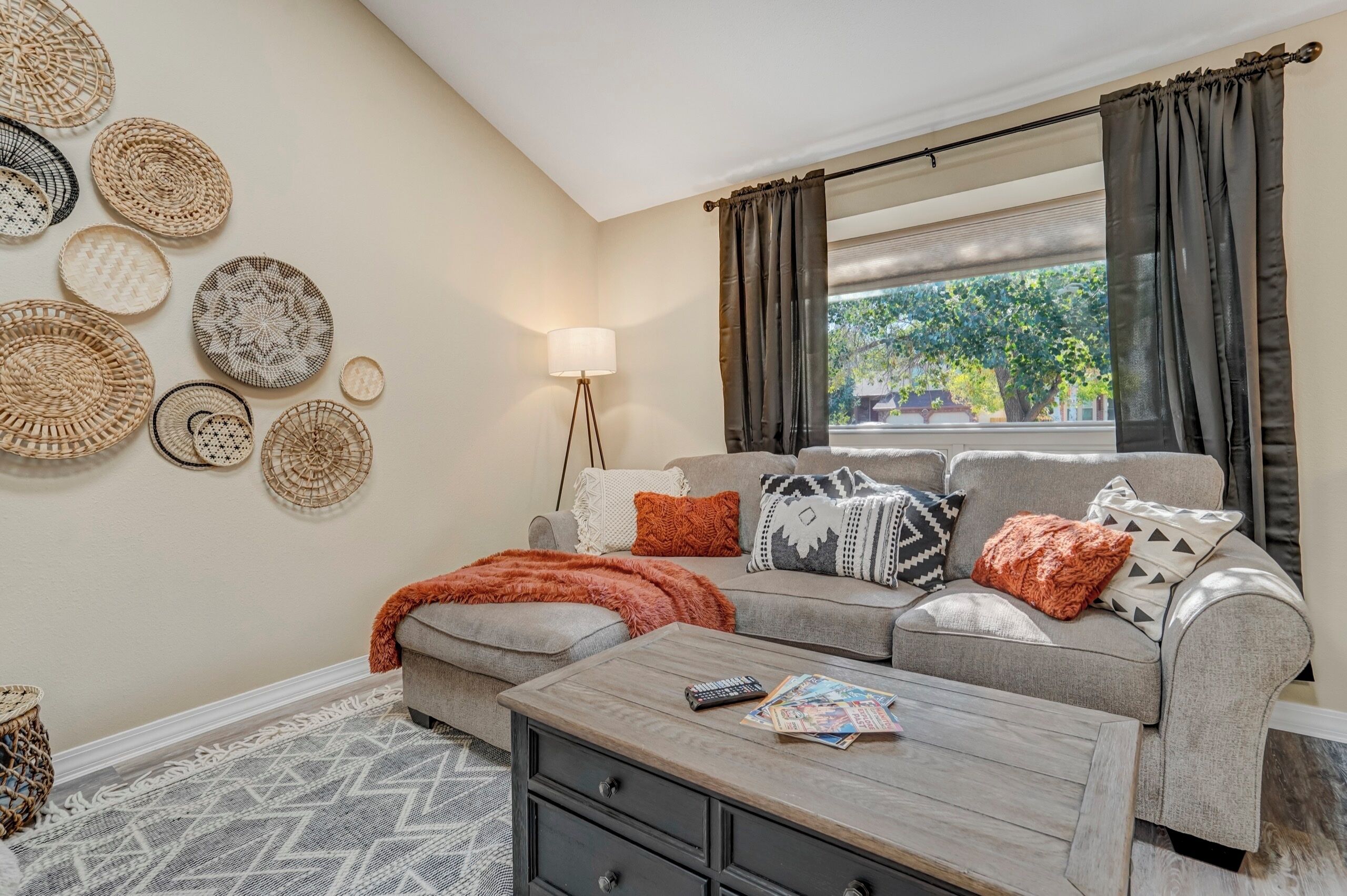 A living room with a sofa, a coffee table, and a lamp. There are woven baskets on the wall and a window with curtains.