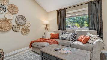 A living room with a sofa, a coffee table, and a lamp. There are woven baskets on the wall and a window with curtains.