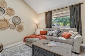 A living room with a sofa, a coffee table, and a lamp. There are woven baskets on the wall and a window with curtains. - Dogs Off-Leash in the Fenced Yard: 4BR Home, Sleeps 9 (Colorado Springs)