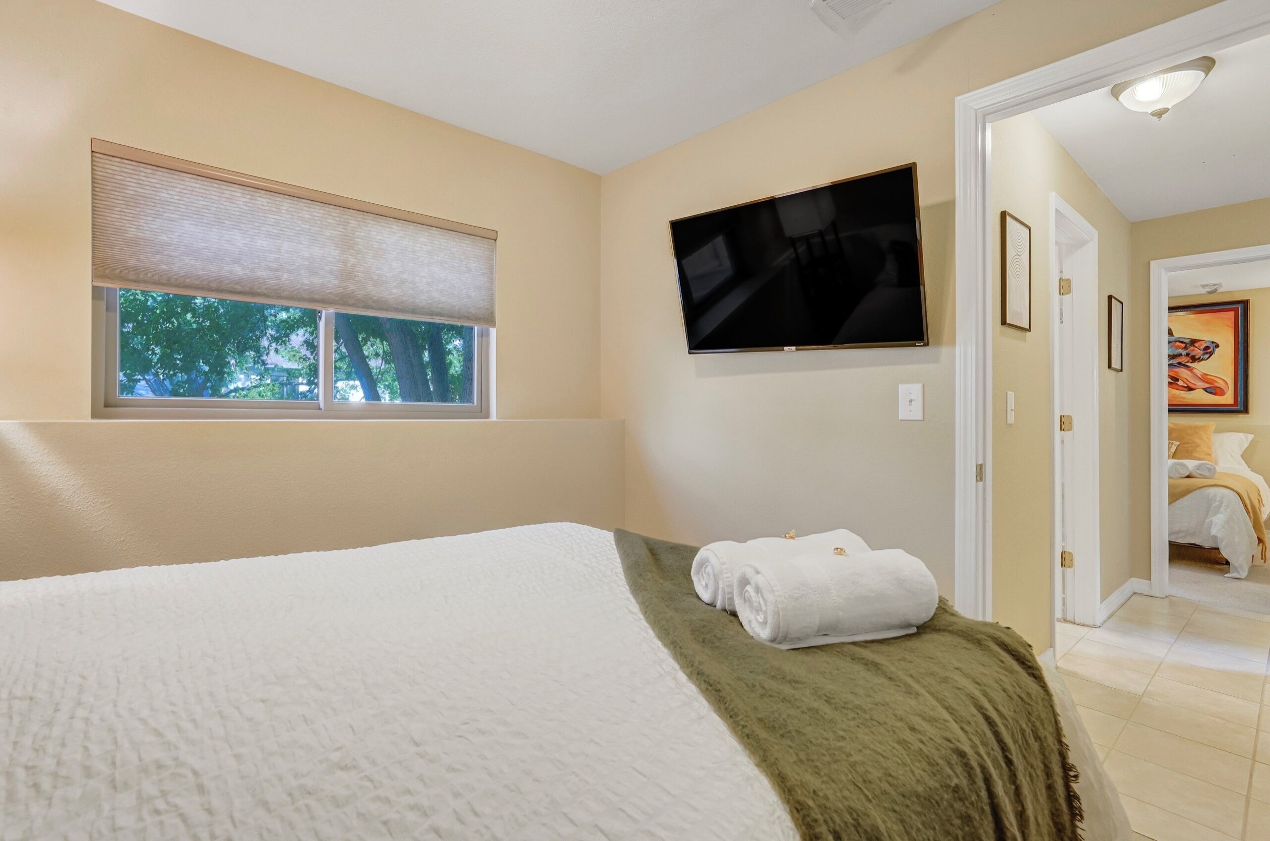 Hotelkamer met een bed, een aan de muur bevestigde televisie, een raam met zonwering en een deur naar een andere kamer.