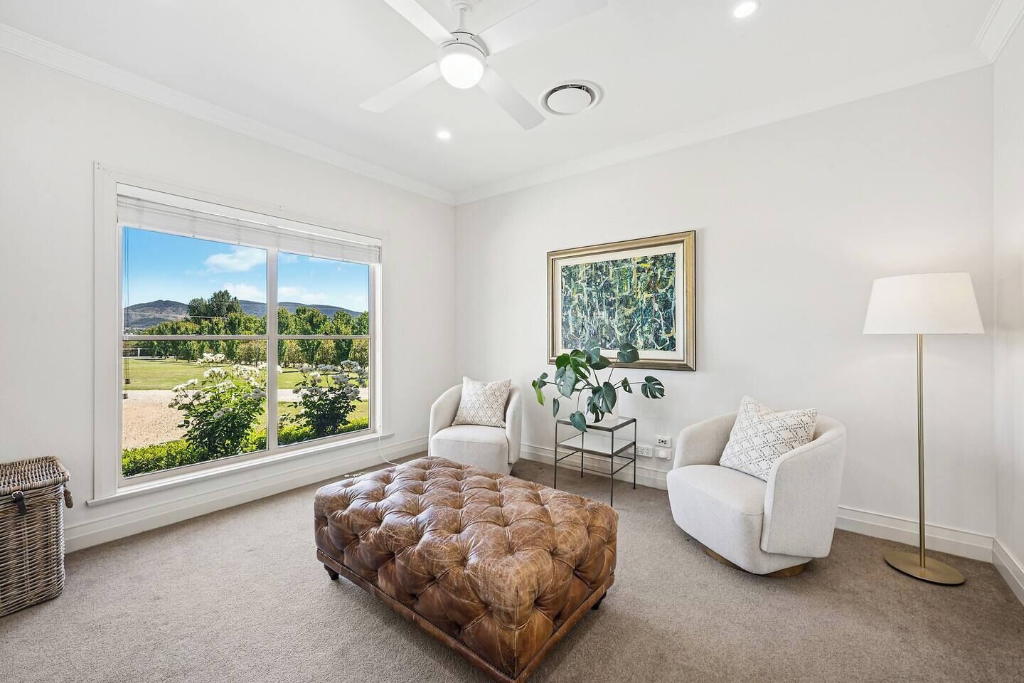A spacious living room with a large window offering a view of a garden and hills. There is a tufted leather ottoman, two white armchairs, and a floor lamp.