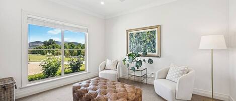 A spacious living room with a large window offering a view of a garden and hills. There is a tufted leather ottoman, two white armchairs, and a floor lamp.