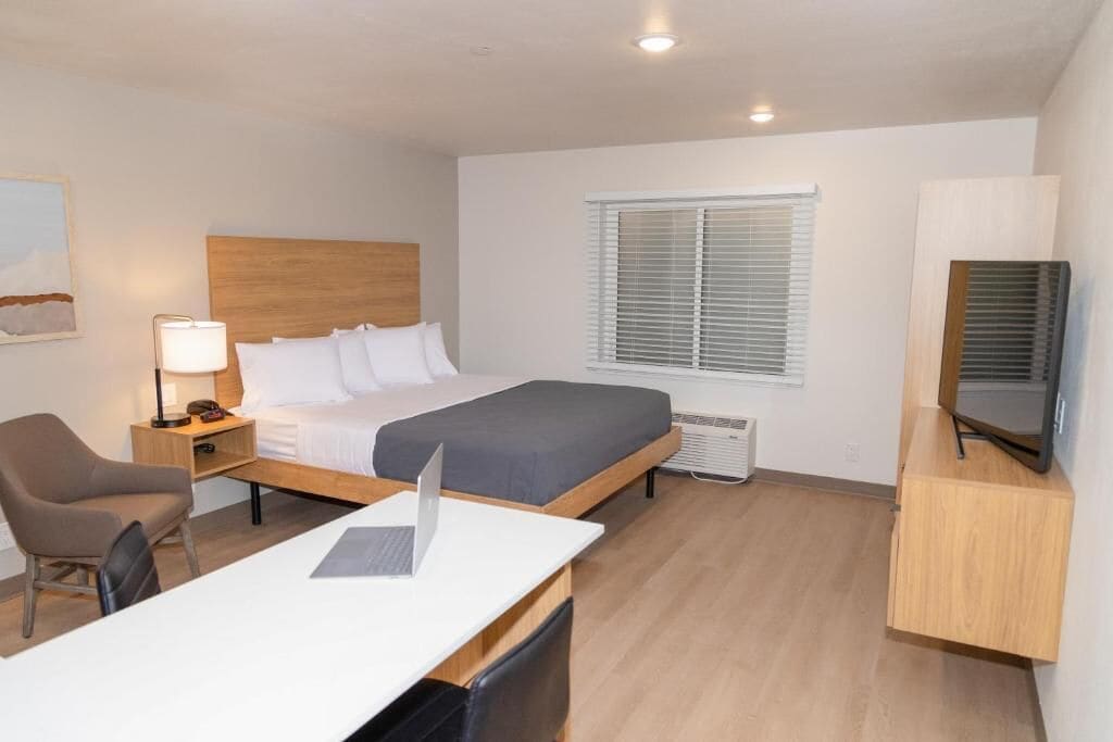 A modern hotel room with a single bed, a desk with a laptop, a chair, a bedside table with a lamp, a TV, and a window with blinds.