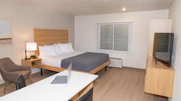 A modern hotel room with a single bed, a desk with a laptop, a chair, a bedside table with a lamp, a TV, and a window with blinds.