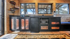 A compact kitchen with a black refrigerator, a microwave, and a built-in oven. There are two windows above the sink and a wooden ceiling. - The Time Machine Treehouse (Rockbridge)