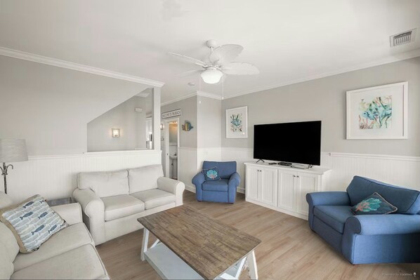 Un salon avec un ventilateur de plafond blanc, un téléviseur à écran plat et une table basse en bois.