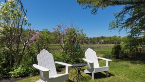 Two white Adirondack chairs with a small round table in a grassy outdoor area with flowering plants and a view of a lake and distant houses. - Rush Pond Retreat at Goose Rocks Beach (Kennebunkport)