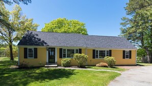 A single-story yellow house with a dark roof, a front porch, and a small front yard with a lawn and shrubs. - Rush Pond Retreat at Goose Rocks Beach (Kennebunkport)