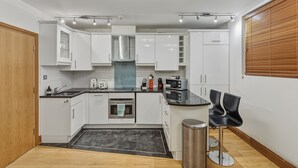 Una cucina moderna con mobili bianchi, una cappa aspirante in acciaio inossidabile, un forno integrato e un piano di lavoro nero. Si nota un piccolo cestino e una sedia nera vicino al bancone.