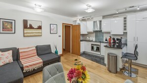 Un appartamento moderno con un divano grigio, un piccolo tavolo da pranzo con un vaso di fiori, una cucina con mobili bianchi e una porta in legno.
