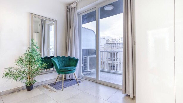 A balcony with a view of buildings, a green chair, a potted plant, and a large mirror. - Central Msida | Modern, Bright & Comfortable (Msida)