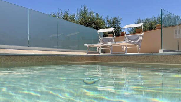 Una piscina con agua cristalina, rodeada por una valla de vidrio y dos tumbonas blancas con toldos.