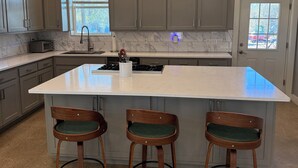 Een keuken met een groot eiland, drie houten barkrukken en uitzicht op de omgeving door een raam.