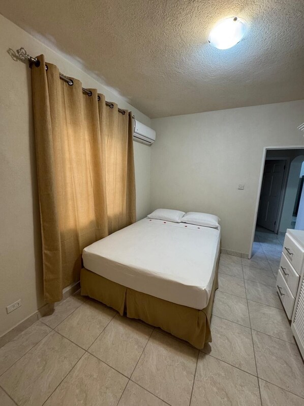 A bedroom with a bed, curtains, an air conditioning unit, and a dresser. - Sakari Ventures Apartments (Bridgetown)