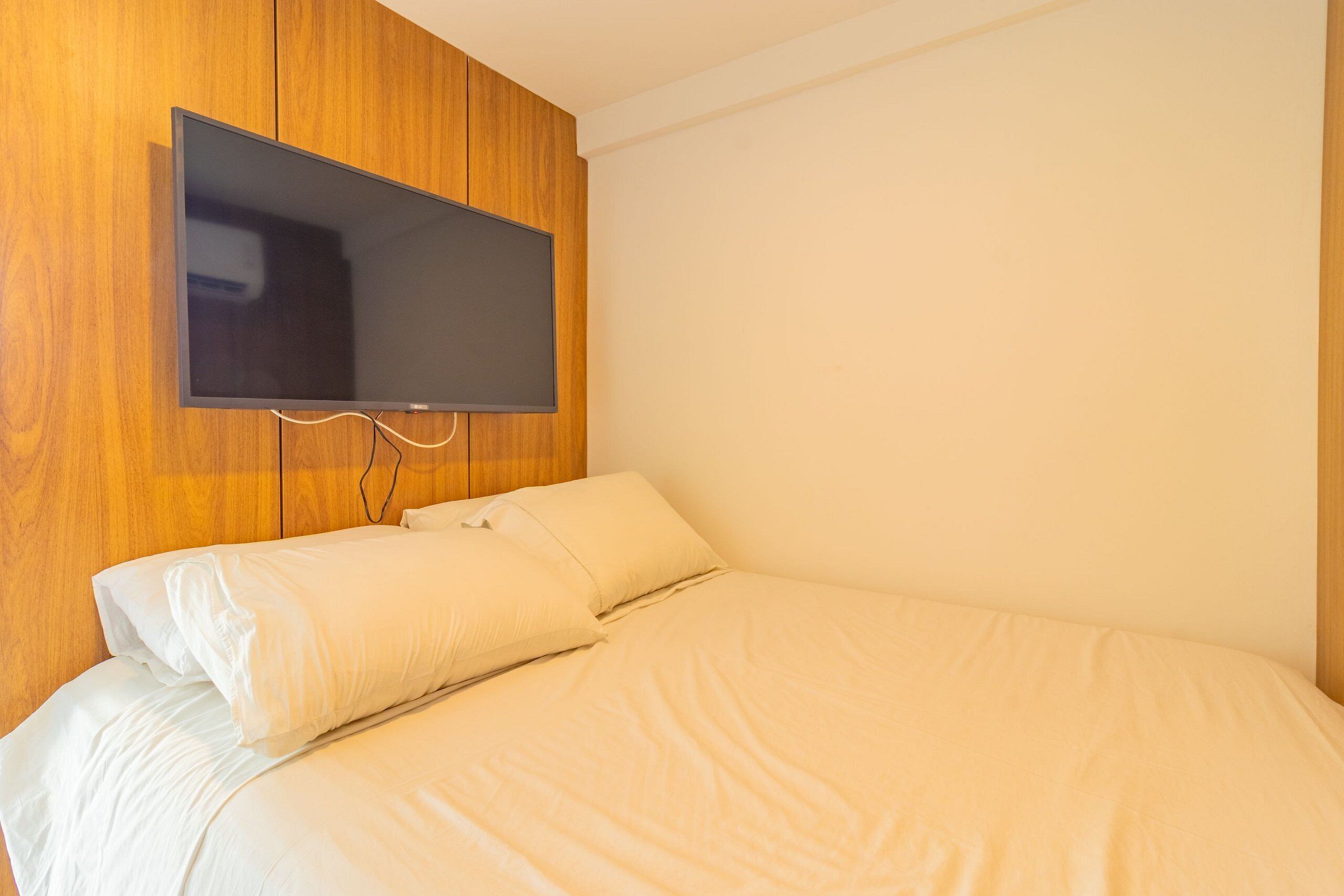 Un dormitorio con una cama, ropa de cama blanca, un televisor de pantalla plana montado y paneles de madera en la pared.