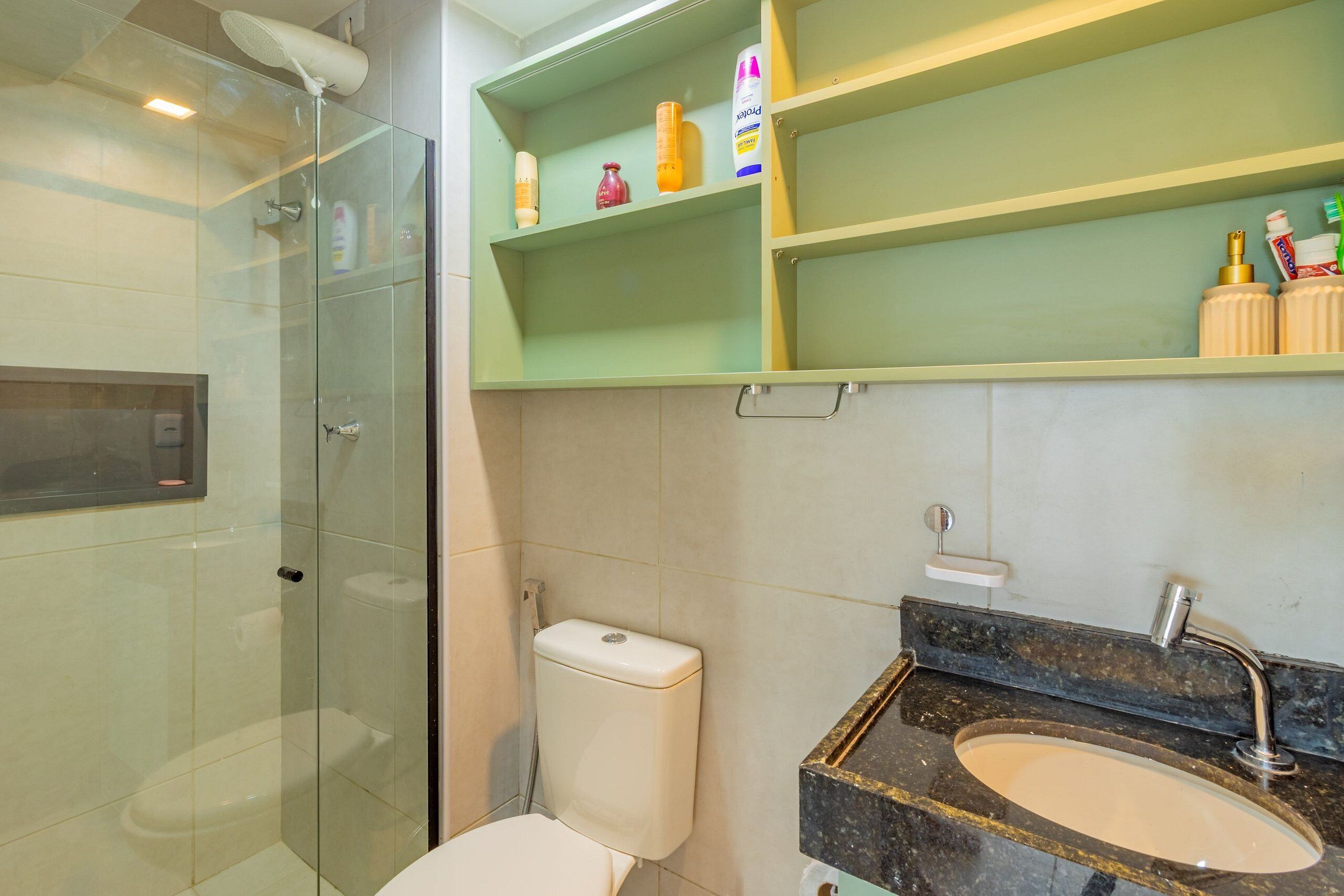 A bathroom with a glass-enclosed shower, a toilet, a sink with a marble countertop, and green shelves with toiletries.