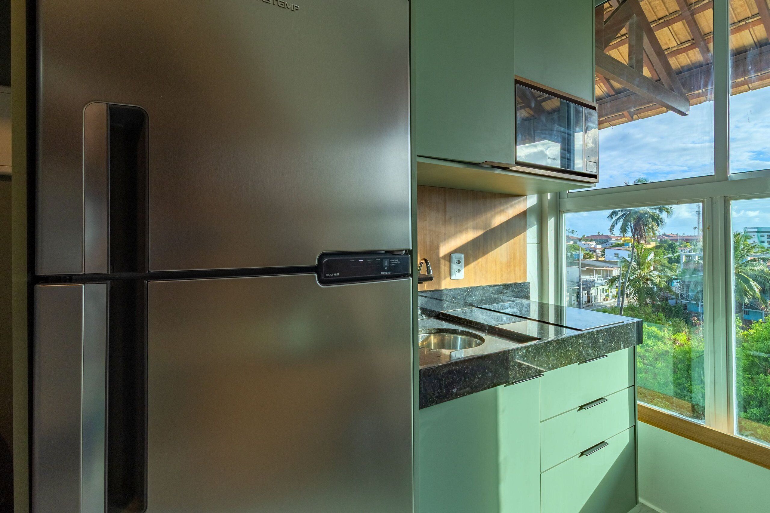 Cozinha com geladeira de aço inoxidável, micro-ondas e janela com vista para palmeiras e edifícios.