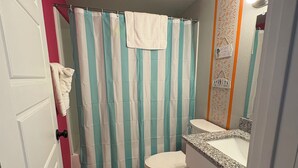 Et badeværelse med blomstret baderomsgardin, toilet, vask med en bordplade i granit og en blomsterdug på gulvet.