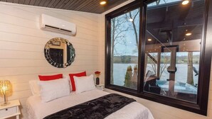 A bedroom with a bed, a nightstand, a lamp, a mirror, and a window with a view of a dock and trees.