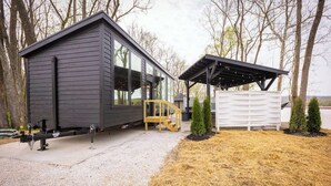 A modern cabin with a large window, a covered outdoor seating area, and a gravel driveway.