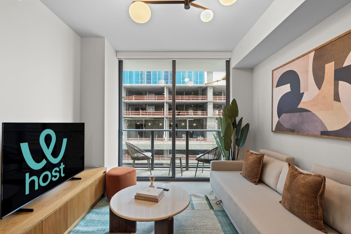 A modern living room with a flat-screen TV, a sofa, a coffee table, and a large window offering a view of a building under construction.