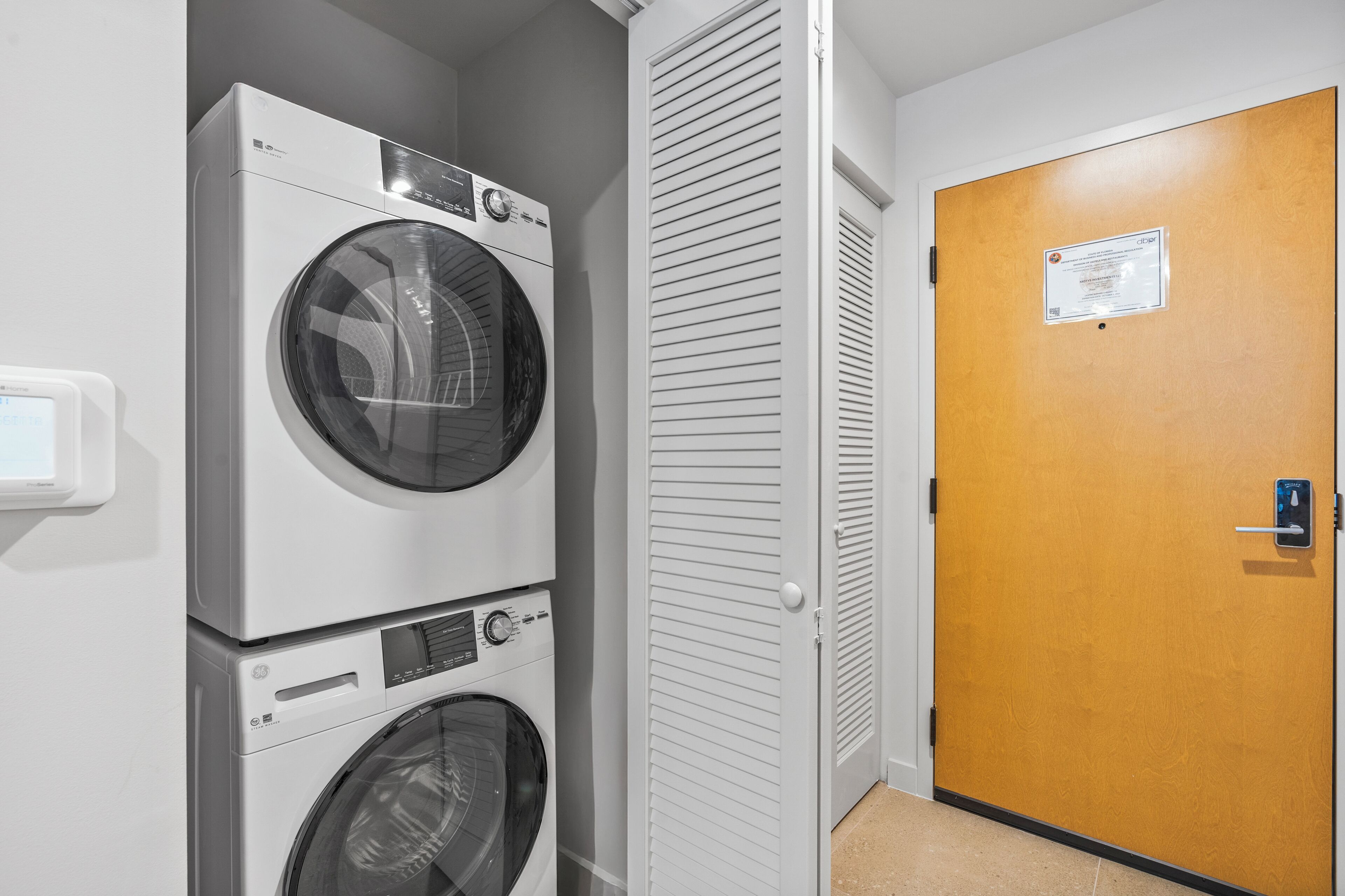 Une buanderie avec des machines à laver et des sèche-linge superposés, une porte blanche avec une pancarte et un sol de couleur beige.