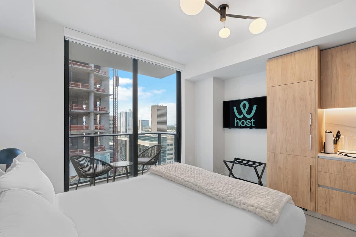A modern hotel room with a large bed, a TV mounted on the wall, a wooden wardrobe, and a balcony with a city view.