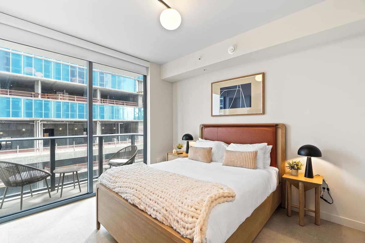 A modern hotel room with a large bed, a bedside table with a lamp, a chair, and a view of a building through large windows.