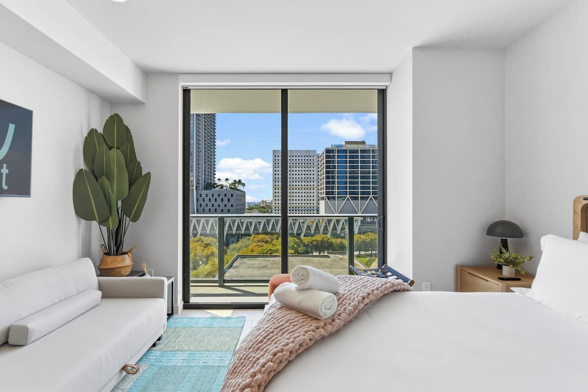 A modern hotel room with a large sliding glass door to the outside, a white sofa, a bed with a pink blanket, and a small bedside table with a lamp and a plant.