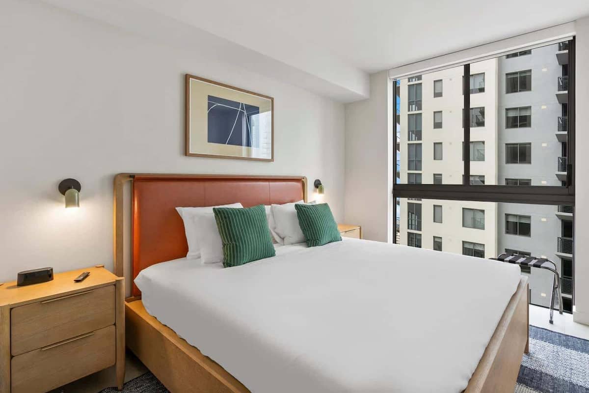 A hotel room with a large bed, a headboard, a nightstand, a remote control, a framed picture on the wall, and a view of the city through a large window.