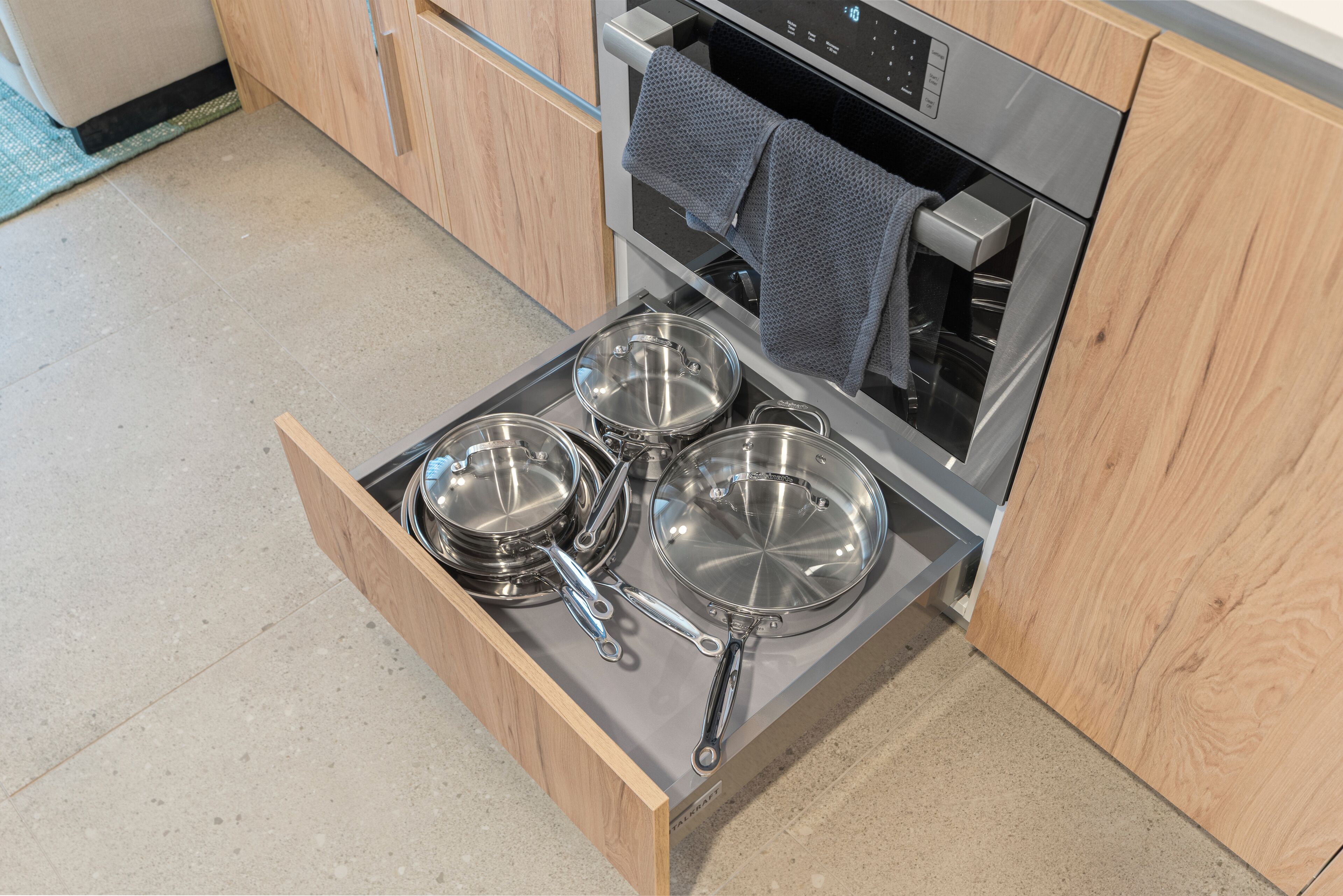A kitchen with a drawer containing stainless steel cookware, a built-in oven, and a towel hanging on the oven door.